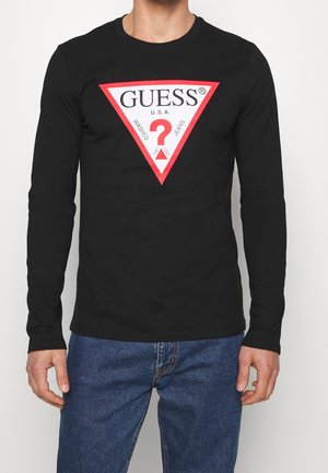 Mężczyzna ubrany w czarną koszulę z długim rękawem z czerwono-białym trójkątnym logo Guess oraz niebieskie jeansy na jednolitym białym tle.