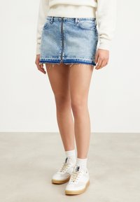 Gonna mini in jeans di colore azzurro chiaro, con orlo sfrangiato, chiusura con zip frontale e due tasche. Abbinata a sneakers bianche e calzini.