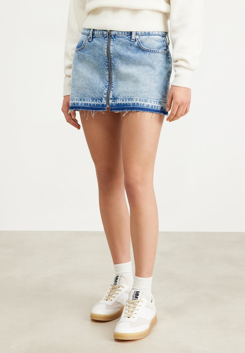 Gonna mini in jeans di colore azzurro chiaro, con orlo sfrangiato, chiusura con zip frontale e due tasche. Abbinata a sneakers bianche e calzini.