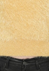 Geel donzig gebreid sweater, cropped boven de taille, dat de bovenkant van zwarte leren broeken met een knoopsluiting laat zien.