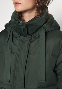 Femme portant une doudoune vert foncé avec un grand col et de larges cordons, ne montrant que la partie inférieure du visage et le haut du torse.