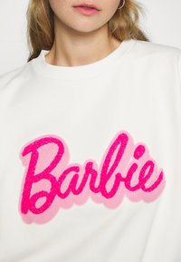 Personne portant un sweat-shirt blanc avec un grand texte "Barbie" rose et magenta sur la poitrine, montrant une partie du cou et des lèvres.