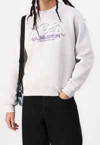 Jasnoszara bluza wykonana z mieszanki bawełny, z połyskującym logo "ALPHA STUDIO EDITION" w iryzujących i purpurowych kolorach. Na sobie czarne spodnie.