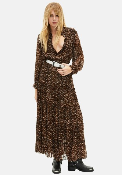 Blonde Frau in einem langen Leopardenmuster-Kleid mit weißem Gürtel und schwarzen Stiefeletten, die vor einem weißen Hintergrund steht.