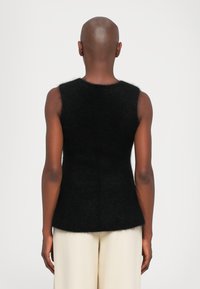 Plikęs žmogus vilki juodą be rankovių pūkuotą viršutinį drabužį ir laisvas grietinėlės spalvos kelnes, matomas iš nugaros ant paprasto fono.