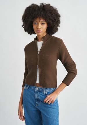 Brun trøje med lynlås og høj krave, der har et tekstureret strikkemønster og diskrete lodrette linjer. Bær den over en lys top, kombineret med blå jeans.