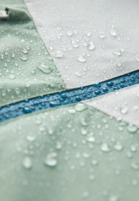 Wasserabweisender Stoff in Hellgrün und Grau, mit einem versteckten blauen Reißverschluss. Die Oberflächenstruktur zeigt Tropfen, die auf wasserfeste Eigenschaften hinweisen.