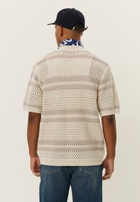 Beiger kurzärmliger Pullover mit Lochmuster und horizontalen Streifen. Verfügt über einen gerippten Kragen und Saum. Kombiniert mit blauen Jeans.