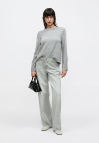 Γυναίκα που φοράει ανοιχτό γκρι πουλόβερ και άνετο ανοιχτό γκρι τζιν, κρατώντας μια μικρή μαύρη τσάντα, στέκεται μπροστά σε λευκό φόντο.