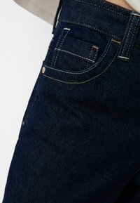 Jeans de mezclilla azul oscuro que presentan una vista lateral con un bolsillo visible, costuras doradas contrastantes y una textura suave.