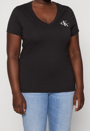 Zwart Calvin Klein T-shirt met V-hals en korte mouwen, gedragen met lichtblauwe spijkerbroek door een staand persoon met ontspannen armen.