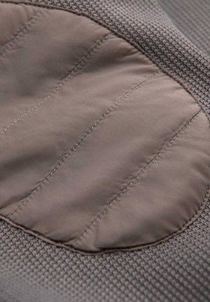 Hellbrauner gepolsterter Stoffflicken auf strukturiertem braunem Strickmaterial mit vertikalen Nahtlinien genäht.