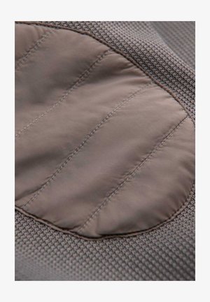 Lys brun polstret stoflap syet på tekstureret brunt strikket materiale med lodrette sømlinjer.