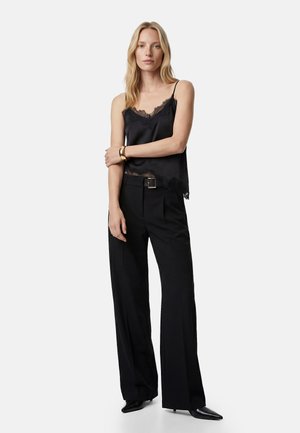 Mujer con camisola negra con encaje, pantalones negros de pierna ancha con cinturón, zapatos negros puntiagudos y pulsera dorada, de pie contra un fondo blanco.