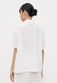 Camisa blanca de manga corta con un patrón de ojales texturizados y diseños florales recortados. Cuello con botones y silueta relajada.