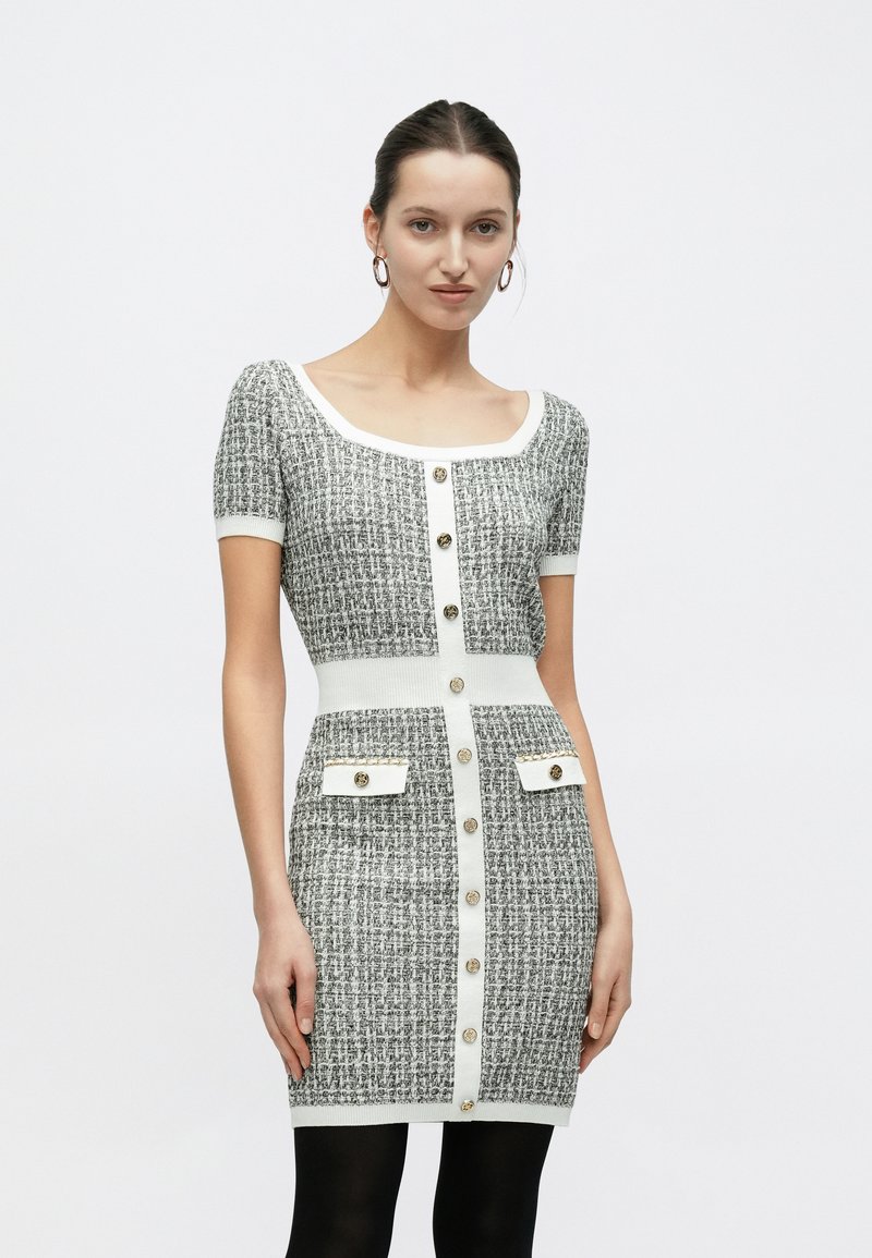 Πλεκτό φόρεμα σε μαύρο και άσπρο με κοντά μανίκια, τετράγωνη λαιμόκοψη, κλείσιμο με κουμπιά μπροστά, δύο τσέπες με καπάκια και λεπτομέρειες σε χρυσό τόνο.