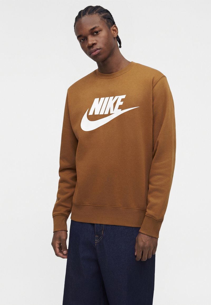 Junger Mann mit geflochtenem Haar, der einen braunen Nike-Sweatshirt mit weißem Logo und dunkelblaue Jeans trägt, steht vor einem schlichten Hintergrund.