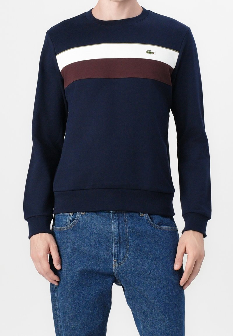 Homme portant un sweat-shirt bleu marine avec des rayures horizontales blanches et marron et un jean bleu, debout devant un fond uni.