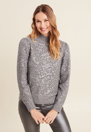 Un pull en tricot gris à col montant, orné de paillettes argentées, avec des poignets et un bas côtelés, porté avec un pantalon gris brillant.