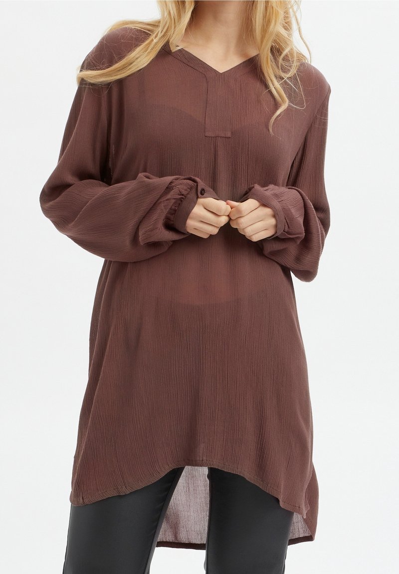 Haut marron à manches longues avec une finition texturée, col en V, détail bouton, et ourlet légèrement incurvé, associé à un pantalon noir.