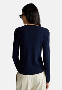 Granatowy sweter z okrągłym dekoltem, długimi rękawami i dopasowanym krojem z prążkowanym wykończeniem. Noszony z jasnymi spodniami.