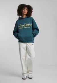 Teal hætteklæd med guld "Prohibited Berlin - New York" tekst, frontlomme, parret med hvide bukser og sneakers, der præsenterer et afslappet look.