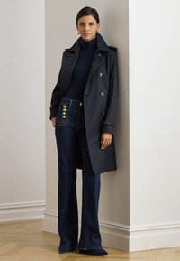 Trench-coat bleu marine à double boutonnage, associé à un jean évasé taille haute avec des boutons dorés et un col roulé sombre.