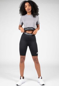 Grijze cropped t-shirt met zwarte Gorilla Wear tailleband, gecombineerd met zwarte fietsbroeken met een wit logo en witte sportschoenen.