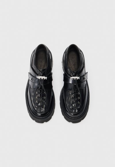 Scarpe in pelle nera con lacci, suole spesse e dettagli in fibbia argentata, abbinate e viste dall'alto su uno sfondo bianco.