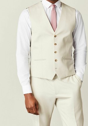 Man wearing cream-colored suit vest and pants with white shirt and pink tie, standing with one hand in pocket.