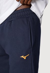 Morske trenirke s elastičnim pojasom, sa bočnim džepovima i narančastim logotipom na lijevom butu. Mekana tekstura tkanine.