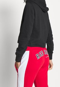 Persona con felpa nera e pantaloni sportivi rossi con strisce bianche sui lati e il testo "RED SOX" sulla schiena, in piedi con una mano in tasca.