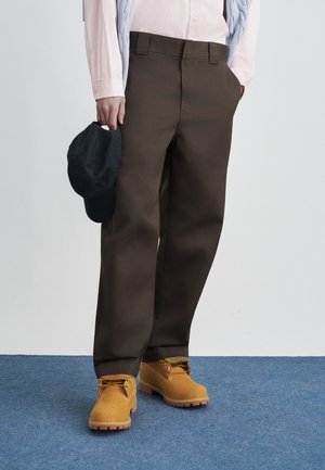 Barna, széles szárú nadrág sima textúrával, sárga csizmával és kézben tartott fekete sapkával párosítva. Világos rózsaszín ing és kék pulóver eldobva.