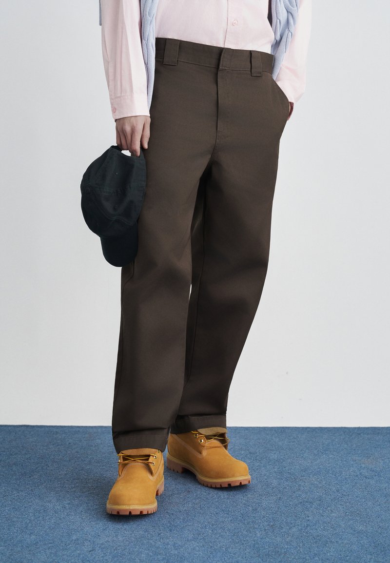 Καφέ παντελόνι με φαρδιά μπατζάκια και λεία υφή, συνδυασμένο με κίτρινες μπότες και μαύρο καπέλο που κρατιέται στο χέρι. Ανοιχτό ροζ πουκάμισο και μπλε πουλόβερ φορεμένα χαλαρά.