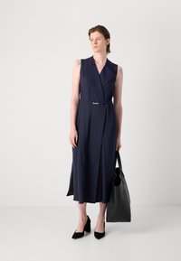 Vestido sin mangas azul marino con rayas finas, ceñido en la cintura y con un escote pronunciado. Combinado con tacones negros y un bolso grande negro.