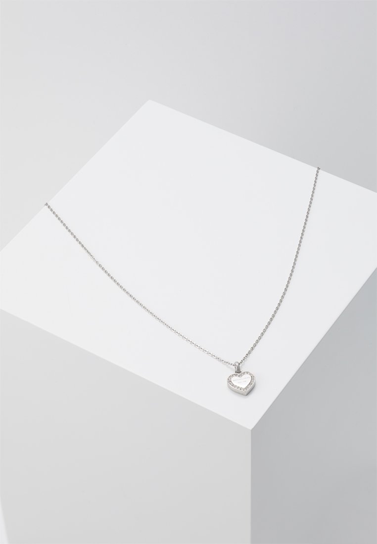 Zarte Silberkette mit einem kleinen herzförmigen Anhänger, der auf einer weißen geometrischen Oberfläche ruht.