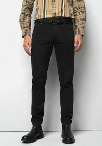 Man in zwarte slim-fit broek, zwarte geweven riem, zwarte leren laarzen en een beige geruite langslijfhemd, staande tegen een witte achtergrond.