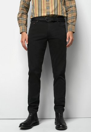 Man wearing black slim-fit pants, black woven belt, black leather boots, and a beige plaid long-sleeve shirt, standing against a white background.