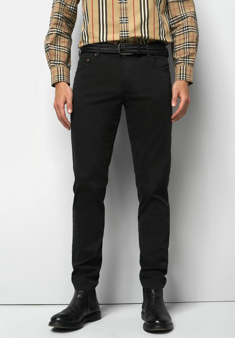 Man in zwarte slim-fit broek, zwarte geweven riem, zwarte leren laarzen en een beige geruite langslijfhemd, staande tegen een witte achtergrond.