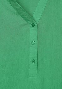 Chemise verte avec un col en V, dotée de trois boutons à l'avant, fabriquée en tissu léger avec une texture lisse.
