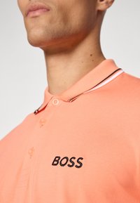 Polo a maniche corte in tessuto pesca con logo "BOSS" ricamato in nero, colletto semplice con righe a contrasto e due bottoni.