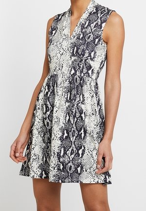 Ärmelloses Kleid mit V-Ausschnitt, schwarz-weißem Schlangenhautmuster, tailliertem Bund und ausgestelltem Rock, der oberhalb der Knie endet, getragen von einer stehenden Person.