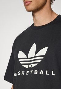 Svart bomullst-shirt med en stor vit adidas-logotyp med tre blad och horisontella ränder, märkt med "BASKETBALL" i feta vita bokstäver.
