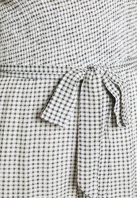 Il tessuto a quadri bianco e nero presenta una vita arricciata con un dettaglio di lacci. La texture è leggera e leggermente sgualcita.
