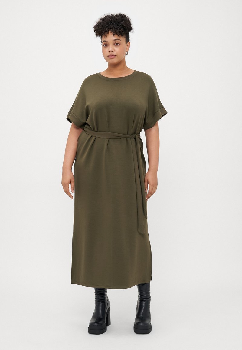 Robe midi vert olive à manches courtes avec une ceinture cravate à la taille, présente une coupe décontractée et une texture douce, associée à des bottes à talons noires chunky.