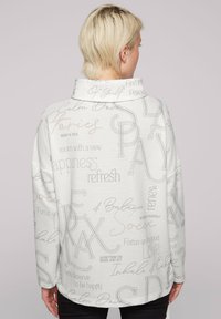 Suéter blanco con patrón y cuello alto, que presenta texto en varias fuentes y colores. Tejido suave y texturizado con un ajuste relajado.