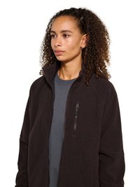 Jeune femme aux cheveux bouclés portant une veste polaire noire zippée sur un t-shirt gris, regardant légèrement vers la gauche sur un fond blanc.