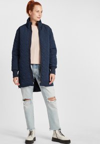 Tengerész kék steppelt hosszú kabát magas gallérral, cipzáras záródással és bordás mandzsettával, világos, koptatott farmernadrággal és fehér fűzős csizmával párosítva.