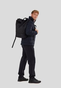 Schwarzer Rucksack mit Roll-Top-Design und verstellbaren Trageriemen, getragen von einer Person in einer dunklen Daunenjacke und schwarzen Jeans, vor einem hellgrauen Hintergrund.