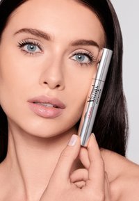 Tubetto di mascara argentato etichettato "Yump! Definition" tenuto vicino al viso; la modella ha occhi azzurri, pelle chiara e labbra rosa chiaro, con capelli lisci e scuri.